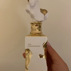 Her Confession Parfym - Lattafa parfymflaska med en unik design. Flaskan är vit med gyllene detaljer och har en skulpturformad topp. Den geometriska basen ger en lyxig känsla. Perfekt för den som vill ha något utöver det vanliga.