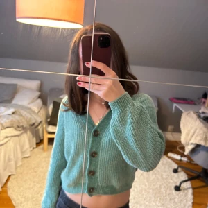 Grön stickad kofta från H&M - Säljer en mysig grön stickad kofta från H&M. Koftan har en croppad passform med knappar framtill och långa ärmar. Perfekt för en avslappnad stil. 💚