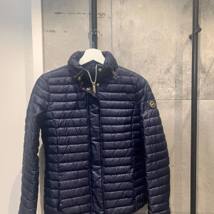 Mörkblå dunjacka från Michael Kors - Snygg mörkblå dunjacka från Michael Kors med quiltad design och hög krage. Jackan har en dragkedja framtill och knappar för extra stil. Perfekt för kyliga dagar och ger en elegant look.