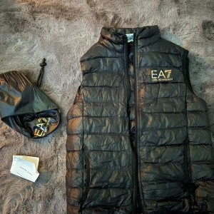Svart dunväst från EA7 - Snygg svart dunväst från EA7 med dragkedja framtill. Västen har ett quiltat mönster och en stilren design. Perfekt för att hålla värmen under kyliga dagar. Kvitto medföljer, Jag är 169 och den passar perfekt på mig.