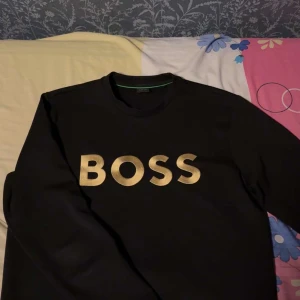 Svart tröja från Boss - Snygg svart tröja från Boss med guldig text på framsidan. Tröjan har långa ärmar och en klassisk rund halsringning. Perfekt för en stilren look.