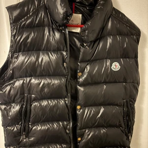 Svart dunväst från Moncler - Snygg svart dunväst från Moncler med dragkedja och knappar. Västen har en hög krage och två fickor med dragkedja. Perfekt för kyliga dagar när du vill hålla stilen.