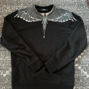 Marcelo burlon - Säljer en svart tröja från Marcelo Burlon med ett coolt vingmönster i grått och silver runt halsen. Tröjan har långa ärmar och en bekväm passform. Perfekt för dig som vill ha en unik stil! Storlek L fast passar perfekt för folk som har storlek M!