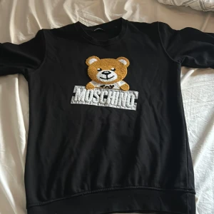 Svart tröja med nallemotiv från Moschino - Säljer en svart tröja från Moschino med ett stort nallemotiv på framsidan. Tröjan har långa ärmar och en rund halsringning. Perfekt för dig som gillar lekfulla och stilrena plagg. 🐻 tröjan är i XL men kan passa som M 