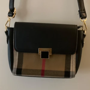 Rutig axelväska med svart lock Burberry - Snygg axelväska med ett klassiskt rutigt mönster i beige, svart och rött. Väskan har ett svart lock och en justerbar axelrem. Den stängs med en elegant fyrkantig spänne och har en dragkedja inuti för extra säkerhet.
