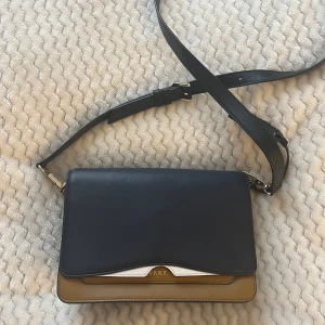 Axelväska från DKNY - Snygg axelväska från DKNY i en stilren design. Väskan har en kombination av svart och beige färg med en justerbar axelrem. Perfekt för att bära dina nödvändigheter med stil.