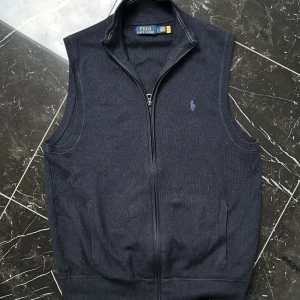 Mörkblå väst från Polo Ralph Lauren - Snygg mörkblå väst från Polo Ralph Lauren med dragkedja och ribbade kanter. Perfekt för lager-på-lager stil. Den har en diskret logga på bröstet. Privat för fler bilder