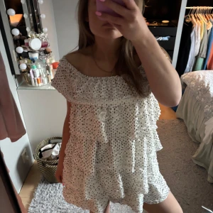 Vit prickig klänning - Säljer en söt vit klänning med prickar och volanger. Klänningen är offshoulder och har en luftig passform, perfekt för varma dagar. Den är kort och har flera lager av volanger.