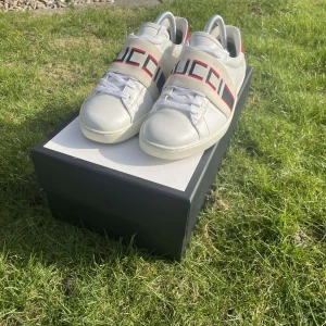 Vita sneakers från Gucci - Säljer ett par stilrena vita sneakers från Gucci med märkets logotyp i rött och grönt på sidan. Skorna har snörning och en klassisk design som passar perfekt för en trendig look. Helt riktiga priset kan diskuteras, säljer åt en kompis box och allt ingår