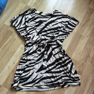 Zebramönstrad klänning med knytband - Snygg zebramönstrad klänning i svart och beige med knytband i midjan. Perfekt för en trendig look. Tillverkad i mjukt polyester för en bekväm passform.