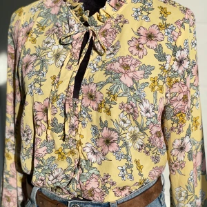 Gina Tricot blommig blus i storlek 40 - Blommig långärmad blus från Gina Tricot i storlek 40. Ljusgul med rosa blommor och knytband fram.  OBS! Den första öglan som håller den översta knappen saknas (se sista bild).