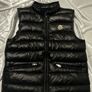 Svart dunväst från Moncler - Snygg svart dunväst från Moncler med dragkedja och två fickor framtill. Perfekt för kyligare dagar med sin quiltade design och stilrena look. Moncler-logga på bröstet ger en exklusiv touch.