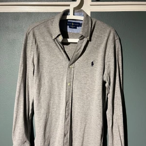 Grå skjorta från Ralph Lauren - Säljer en stilren grå långärmad skjorta från Ralph Lauren. Skjortan har knappar framtill och en broderad logga på bröstet. Perfekt för en klassisk look.