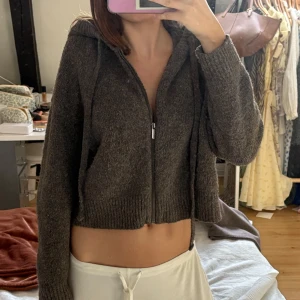Brun stickad hoodie med dragkedja - Säljer en mysig brun stickad hoodie med dragkedja framtill. Tröjan är croppad och har långa ärmar samt en huva med snören. Perfekt för kyligare dagar!