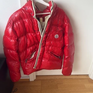 Röd dunjacka från Moncler - Säljer en snygg röd dunjacka från Moncler med dragkedja och två praktiska fickor framtill. Jackan har en hög krage och Moncler-loggan på bröstet. Perfekt för kyliga dagar!