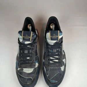 Blåa Valentino rockrunners - Snygga Valentino Garavani sneakers med ett coolt kamouflagemönster i blå och grå toner. Skorna har en unik design med detaljer i mocka och en kontrasterande röd häl. Perfekta för att ge din outfit en edgy touch. Skorna skickad med og dustbag men utan og box