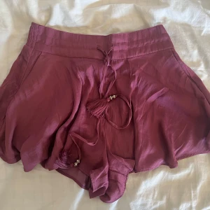 Rosa shorts med knytband - Superfina luftiga shorts i en mörkrosa nyans med volangig nederkant och dekorativt knytband med tofsar och pärlor i midjan. har en flowy passform och är tillverkad i ett mjukt, lätt tyg. Har ett smock på bakdelen och shortsen är högmidjade. Priset går att diskuteras :) 