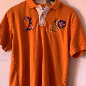 Orange pikétröja med brodyr och tryck - Orange pikétröja från Polo Club England med korta ärmar, klassisk krage och vit knappslå. Broderad logga, stort siffratryck och patch med brittisk flagga på bröstet. Perfekt för dig som gillar sportig stil.