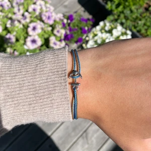Blått och brunt armband med ankare  - Dubbeltrådigt armband i blått och brunt med en liten silvrigt ankare. Justerbar storlek med knutar och enkel, stilren design som passar perfekt till en avslappnad look. Ankaret i metall ger en cool detalj till armbandet.