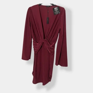 Vinröd kortklänning från NLY One - Supersnygg vinröd kortklänning från NLY One med lång ärm, djup v-ringning och cool knutdetalj framtill i midjan. Klänningen har en figurnära passform och är gjord i ett mjukt, stretchigt material som känns bekvämt mot huden.