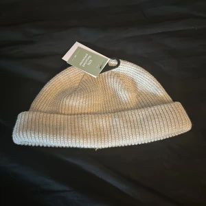 Beige ribbad stickad mössa - Snygg beige stickad mössa med bred uppvikt kant och ribbad struktur. Perfekt accessoar för kyliga dagar och enkel att matcha med olika outfits. Stilren och tidlös design som passar de flesta.