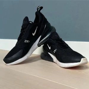 Nike Air Max 270 i svart och vitt - Säljer ett par svarta Nike Air Max 270 sneakers med vita detaljer och stor synlig Air-enhet i hälen. Skorna har mesh-ovandel, snörning och en sportig vibe. Perfekta för dig som gillar streetstyle och bekväma sneakers. Finns dock två smål hå vid skorna med syns knappt.