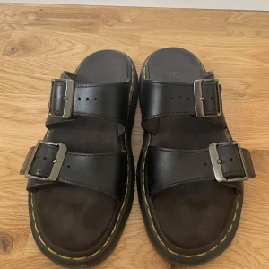 Svarta Dr. Martens sandaler med spännen - Chunky svarta sandaler från Dr. Martens med två breda remmar i skinn och stora metallspännen. Klassisk gul söm runt sulan och platt sula med öppen tå. Perfekta för en edgy och avslappnad stil.