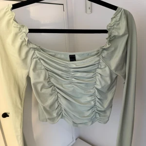 Ljusgrön långärmad topp med rynk - Supersöt ljusgrön långärmad topp från H&M med rynkade detaljer framtill och vid axlarna. Toppen har en bred, något fyrkantig urringning och är figurnära i modellen. Perfekt för dig som gillar trendiga och feminina plagg.