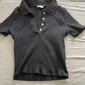 Svart ribbad pikétröja från Zara - Svart ribbad pikétröja från Zara med klassisk krage och fyra vita knappar framtill. Tröjan har korta ärmar och är tillverkad i ett mjukt bomullsmaterial. Perfekt för en clean och stilren look.
