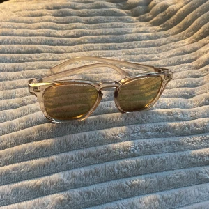 Solglasögon från Le Specs - Snygga solglasögon från Le Specs med transparent båge och guld-brun glas. Klassisk wayfarer-inspirerad form som ger en cool vibe. Lätta och bekväma att bära, perfekta för soliga dagar. Passar dig som gillar stilrena accessoarer med modern touch. De har repor som är synliga i ett viss ljus och även en svart fläck på högra glaset i hörnet. Har inget fodral.