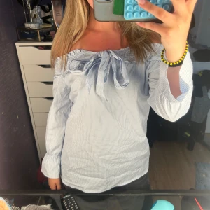 Offshoulder blus med knyt och ränder - Säljer en ljusblå och vit randig offshoulder-blus med smockad kant upptill och stor knytning framtill. Blusen har trekvarts ärmar med lätt puff och avslappnad passform. Perfekt för dig som gillar en söt och trendig stil.