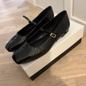 Svarta lackade ballerinaskor med spänne - Stilrena svarta ballerinaskor i glansigt lackmaterial med fyrkantig tå och smal rem över foten med guldfärgat spänne. Perfekta för en chic och trendig look. Storlek 39