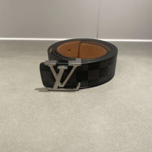 Louis Vuitton rutig svart skinnbälte - Snyggt svart bälte från Louis Vuitton med klassiskt rutigt mönster och stor LV-logga i silver på spännet. Bältet är gjort i exklusivt skinn med brun insida och har en stilren, modern look som sticker ut.