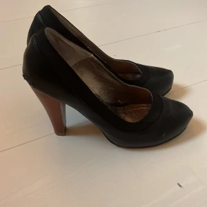 Svarta pumps med klack, storlek 38 - Klassiska svarta pumps med spetsig tå och hög klack. Skorna har en snygg rundad öppning och insida i metallicfärg. Perfekta för dig som vill ha en stilren och tidlös look. Klacken är i en kontrasterande brun nyans.