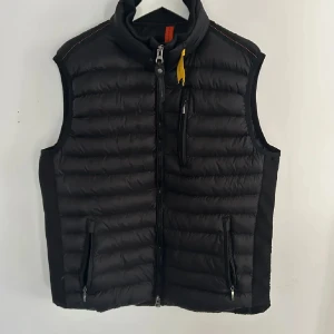 Svart dunväst från Parajumpers XL - Svart dunväst från Parajumpers med quiltad design, hög krage och dragkedja framtill. Västen har tre fickor med dragkedja och en gul detalj vid halsen. Perfekt för lager-på-lager och snygg till både hoodie och t-shirt.