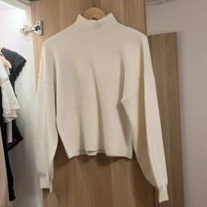 Vit polotröja med hög krage - En stilren vit polotröja med hög ribbad krage och långa ärmar. Tröjan har en rak passform och är tillverkad i ett mjukt material som känns skönt mot huden. Perfekt för lager-på-lager och enkel att matcha med olika outfits. Aldirg använd