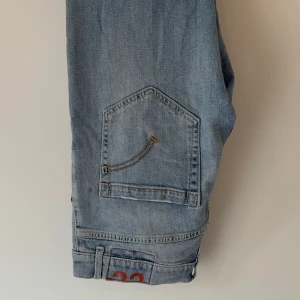 Ljusblå jeansbyxor med orange detalj - Säljer ett par ljusblå jeans från Dondup med raka ben och klassisk femficksdesign. Snygg broderad detalj på bakfickorna och en orange 33-detalj vid benslutet. Jeansen har normal passform och är tillverkade i bomull. W31