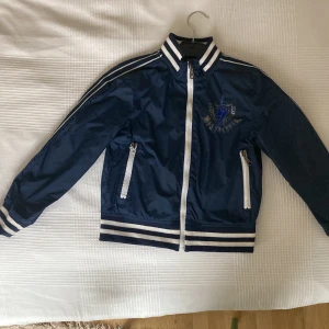 Blå bomberjacka från Replay, 130cm - Snygg mörkblå bomberjacka från Replay med vita ränder på krage, ärmslut och midja. Jackan har dragkedja framtill, två fickor med dragkedja och coolt blixt-tryck på bröstet. Perfekt för dig som vill ha en sportig och trendig look.