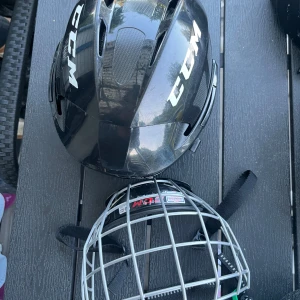 Svart hockeyhjälm från CCM - Säljer en svart hockeyhjälm från CCM med tillhörande galler. Hjälmen har ventilerande hål och justerbara remmar för perfekt passform. Gallret är i silverfärgad metall och skyddar ansiktet under match och träning.