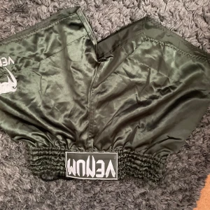 Gröna Venum träningsshorts XXL - Snygga gröna träningsshorts från Venum i glansigt syntetmaterial. De har bred resår i midjan med Venum-logga framtill och coolt tryck på sidan. Perfekta för dig som gillar kampsport eller gymmet.