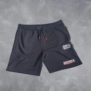 Hoodrich shorts - Svarta shorts från Hoodrich med vit och röd logga på ena benet och patch-detalj. Mjukt material, elastisk midja med snörning och röda detaljer på snöret. Perfekta för chill eller träning.