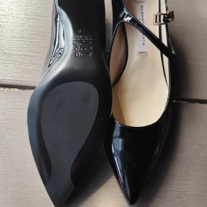 Svarta lackpumps med spetsig tå - Snygga svarta pumps i lack från Roberta Festa med elegant spetsig tå och smal rem med guldfärgat spänne över foten. Perfekta för dig som gillar klassisk och stilren design med en modern twist.