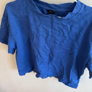 Blå croppad t-shirt från Lindex - En enkel och stilren blå croppad t-shirt från Lindex. T-shirten har rund halsringning och korta ärmar, perfekt för en avslappnad look. Tillverkad i mjuk bomull som känns skön mot huden.
