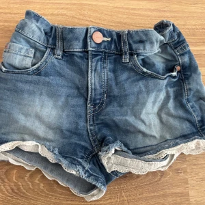 Blå jeansshorts med spetskant - Snygga blå jeansshorts från Narrow i klassisk denim med coola fickor och vit spetskant längst ner. Shortsen har normal passform och knappstängning framtill. Perfekta för varma dagar när du vill ha en chill och trendig look.