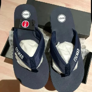 Mörkblå sandaler från italienska Colmar - Snygga mörkblå sandaler från Colmar med vit logga på remmen och rund tå. Sköna flip-flop-modell med textilrem och platt sula, perfekta för varma dagar. Klassisk och stilren design med vit rand längs sulan.