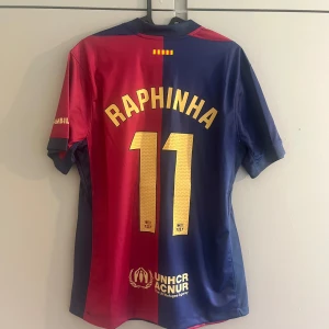FC Barcelona Raphinha 11 matchtröja Nike - FC Barcelona matchtröja med Raphinha 11 på ryggen. Klassisk halva-röd halva-blå design, officiella klubbmärket och Nike-logga framtill, samt sponsorer som Spotify och Ambilight tv. Tillverkad i lätt och ventilerande polyester, perfekt för fotbollsälskare.