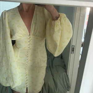 Gul blommig Florence-klänning från Adoore - Supersöt kortklänning från Adoore i ljusgul nyans med små blommor. Djup v-ringning, markerad midja och klädda knappar framtill. Vida, långa ärmar med resår vid handlederna. Perfekt för sommarens alla tillfällen!