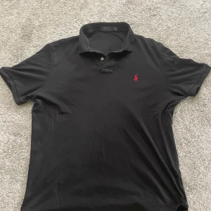 Svart pikétröja från Polo Ralph Lauren - Klassisk svart pikétröja från Polo Ralph Lauren i bomull, med röd broderad logga på bröstet. Tröjan har korta ärmar, krage och två knappar framtill. Perfekt för en stilren och avslappnad look.