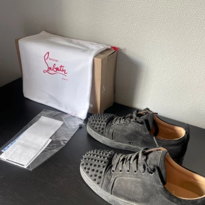 Svarta sneakers från Christian Louboutin - Svarta sneakers i mocka från Christian Louboutin med coola nitar över tån. Skorna har snörning och en platt sula, vilket ger en edgy vibe. Perfekta för dig som vill sticka ut med en lyxig och unik detalj i din outfit.