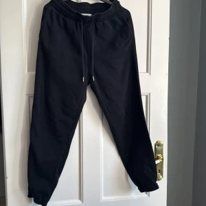 Svarta mjukisbyxor från H&M, storlek M - Svarta mjukisbyxor från H&M i storlek M. Byxorna har resår i midjan med snörning och muddar vid bensluten. Tillverkade i en mjuk bomullsblandning, perfekta för chill dagar eller när du vill vara bekväm. Klassisk och enkel design utan tryck eller mönster.